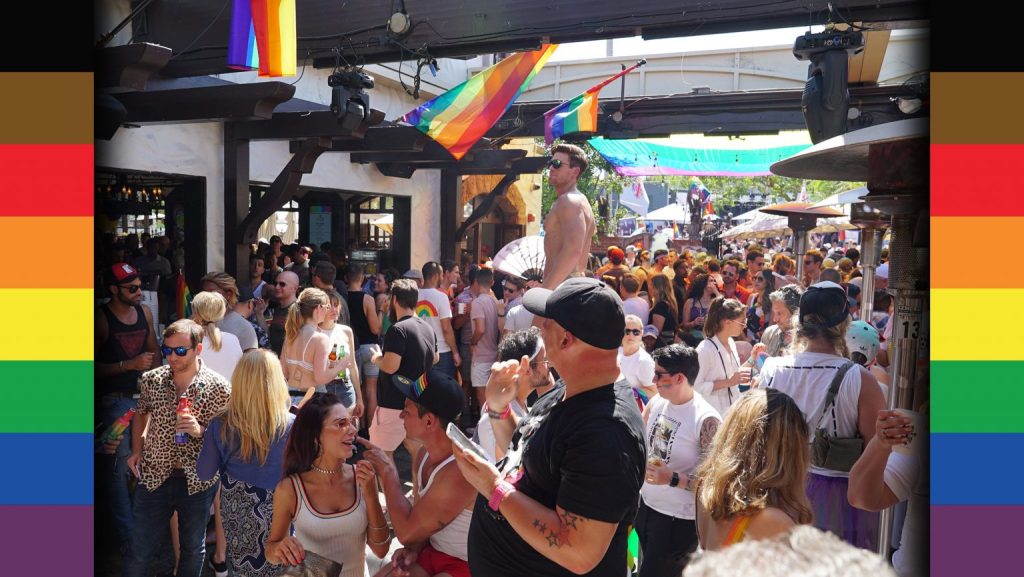 a crowd of people under the summer sun in the courtyard of a gay bar bedecked by pride flags